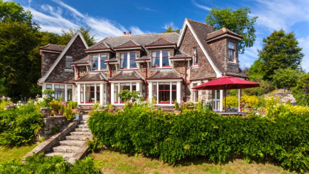 Large two-story house with stone and wood exterior, reminiscent of a Jane Austen novel, surrounded by lush greenery, gardens, and a red patio umbrella under a clear blue sky.