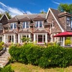 Large two-story house with stone and wood exterior, reminiscent of a Jane Austen novel, surrounded by lush greenery, gardens, and a red patio umbrella under a clear blue sky.