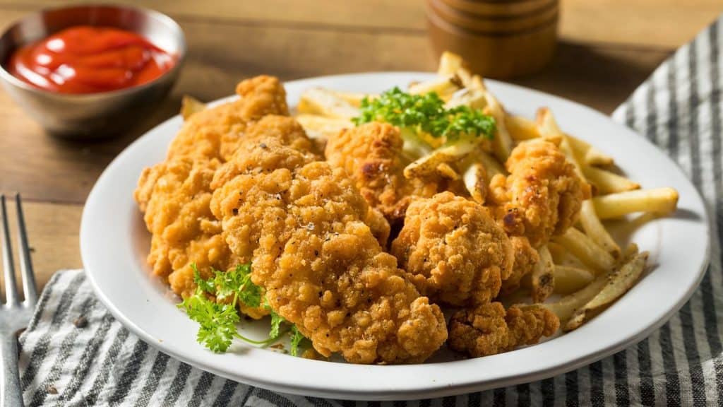 A plate of fried chicken tenders and French fries garnished with parsley, with a bowl of ketchup in the background.