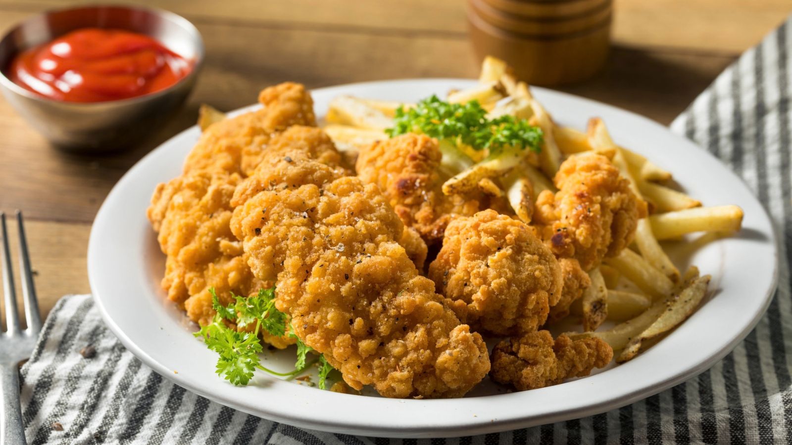 A plate of fried chicken tenders and French fries garnished with parsley, with a bowl of ketchup in the background.