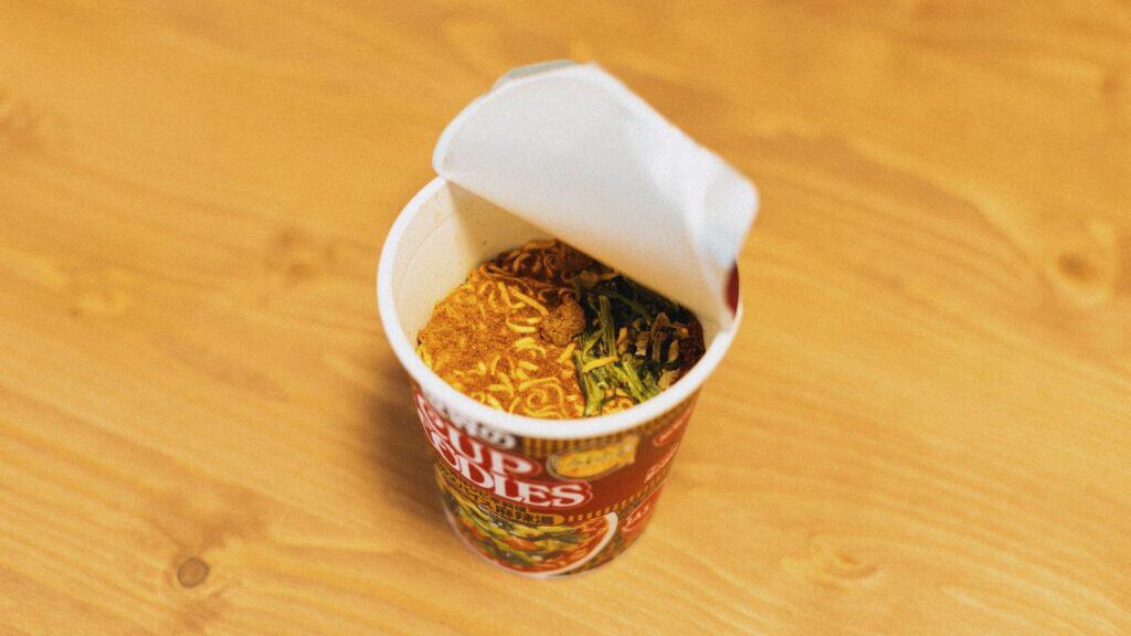 An open cup of instant noodles with seasoning and dried vegetables on a wooden surface, these essential dorm pantry staples offer a quick and satisfying meal anytime.