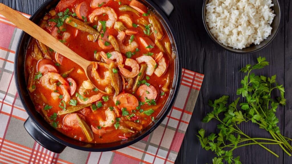 A pot of shrimp and vegetable stew with a wooden spoon, placed on a plaid cloth, highlights the flavors of fall vegetables coming into season, next to a bowl of white rice and a bunch of fresh parsley.