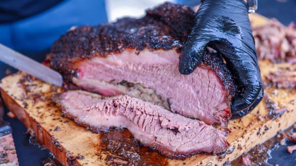 A gloved hand slices a large piece of smoked brisket on a wooden cutting board, revealing a pink interior and dark crust—just the kind of mouthwatering scene you’d find at a Longhorn tailgates brisket contest.