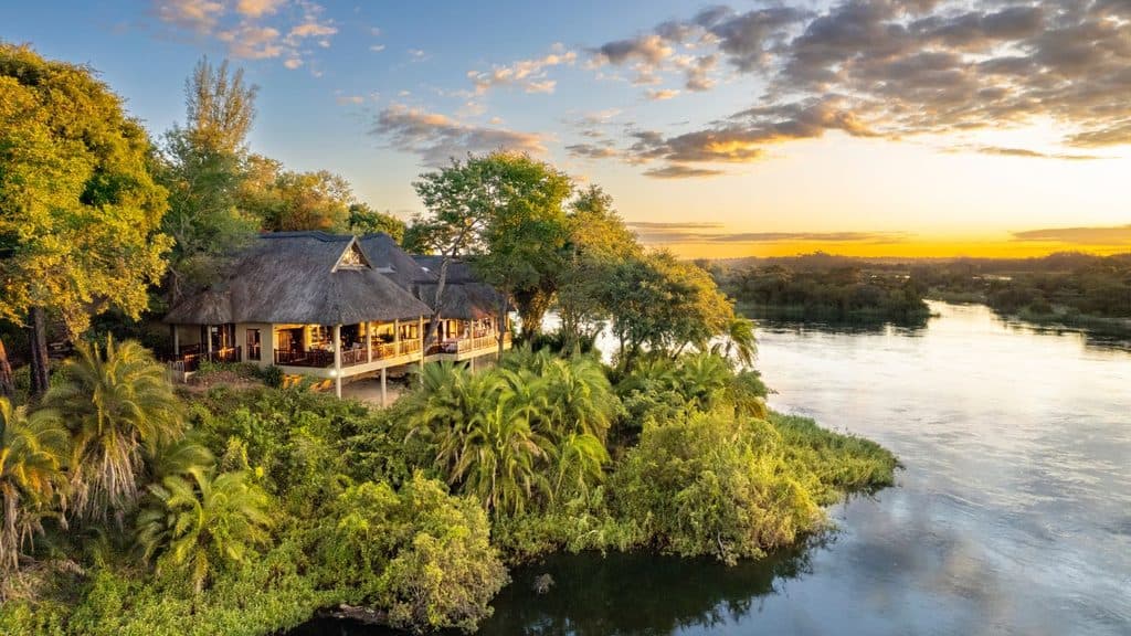 A thatched-roof lodge sits among lush greenery on the edge of a river at sunset, with trees and clouds in the background.