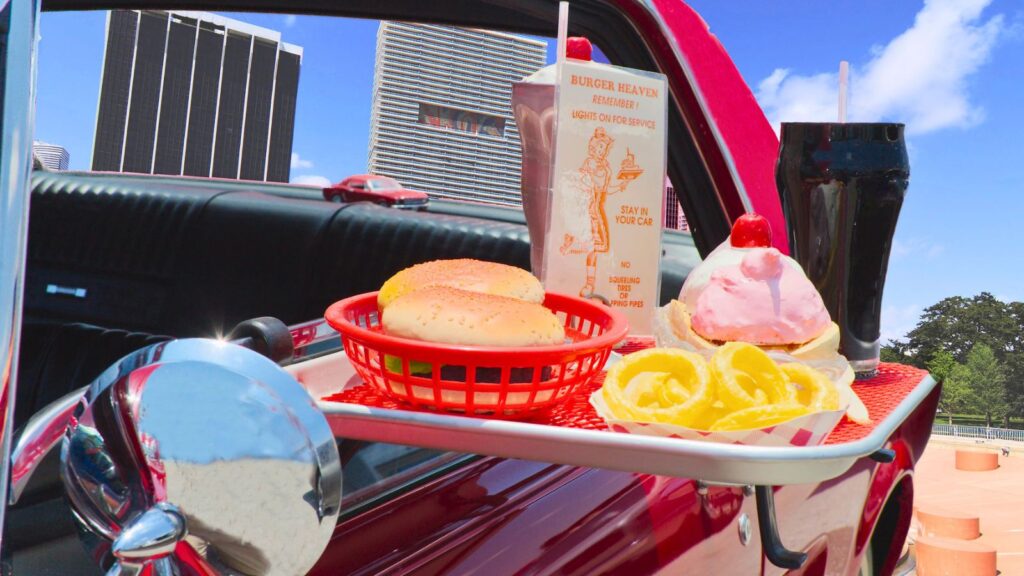 A carhop tray attached to a red car holds burgers, onion rings, a drink, and a pink ice cream sundae, just the way drive-in carhop regulars remember, with city buildings visible in the background.