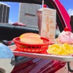 A carhop tray attached to a red car holds burgers, onion rings, a drink, and a pink ice cream sundae, just the way drive-in carhop regulars remember, with city buildings visible in the background.