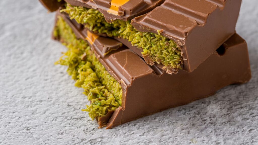 Close-up of stacked milk chocolate bars with a green pistachio filling, placed on a textured gray surface.