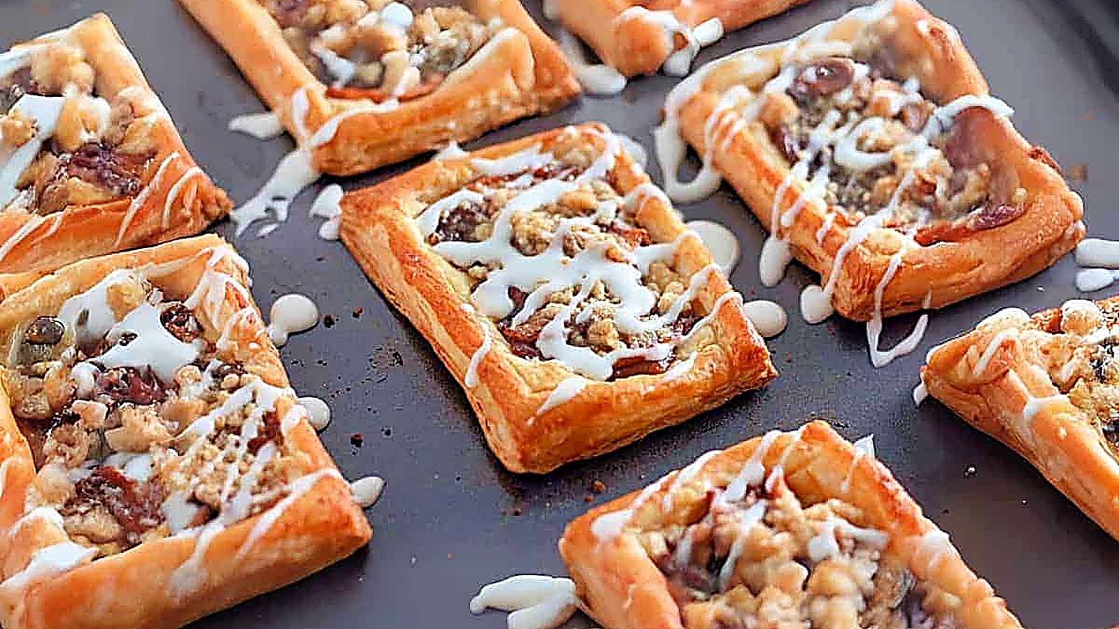 Golden brown Dutch Apple Danishes, topped with a fruit and nut filling, drizzled with white icing, displayed on a dark baking sheet.
