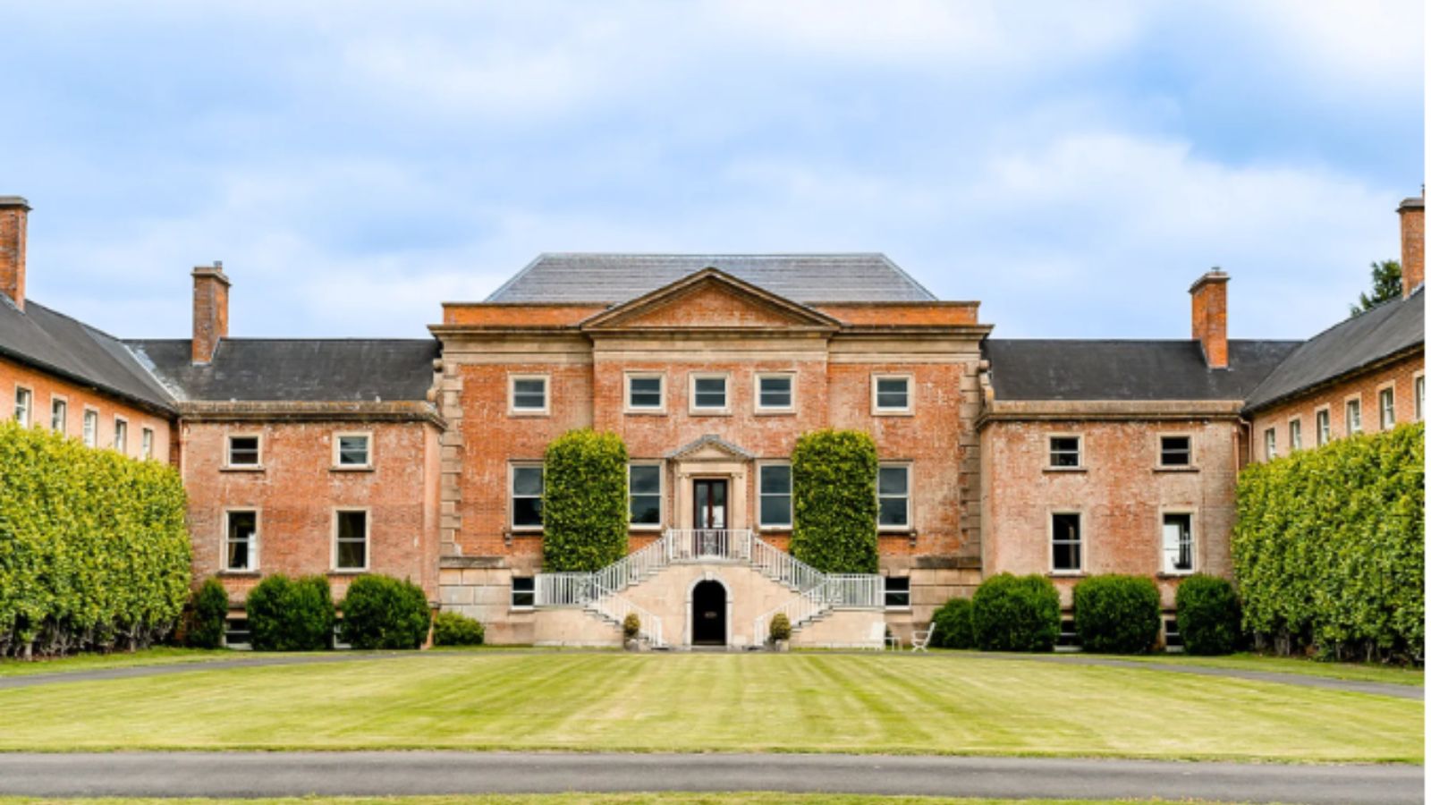 Large red-brick mansion with symmetrical wings, central entrance with stairs, manicured lawn, and trimmed hedges under a partly cloudy sky.