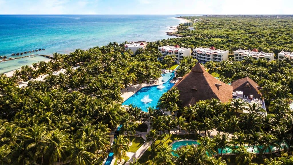 Aerial view of a tropical beachfront resort with a large thatched-roof building, swimming pools, palm trees, and white buildings near the ocean.