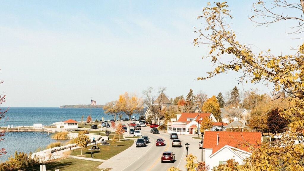 Experience quintessential fall travel in this charming small lakeside town, where cars cruise along the road past red-roofed buildings and vibrant autumn trees. An American flag waves proudly while a pier stretches into the water beneath a clear sky&mdash;perfect for those who love exploring small towns in the heart of autumn.