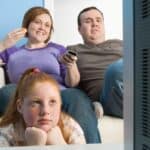 A young girl sits in front of a couch, resting her chin on her hands, while two adults behind her watch 80s and 90s must-see TV and eat snacks.