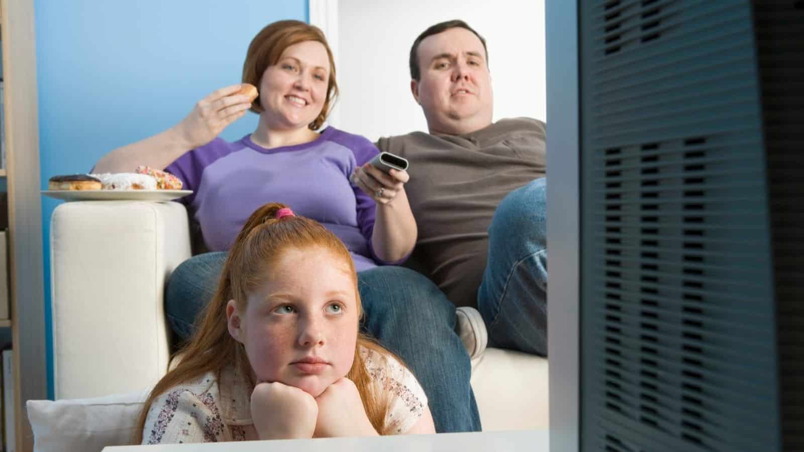 A young girl sits in front of a couch, resting her chin on her hands, while two adults behind her watch 80s and 90s must-see TV and eat snacks.