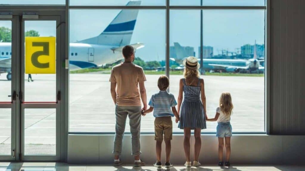 A family of four stands inside an airport terminal, holding hands and looking out the window at airplanes on the runway, perhaps anticipating potential US airports flight delays as they watch.