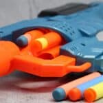 Close-up of a blue and orange toy dart gun with foam darts in the chamber and more foam darts lying beside it on a gray surface&mdash;one of the latest toy fads that&rsquo;s sure to be at the top of every kid&rsquo;s wish list.