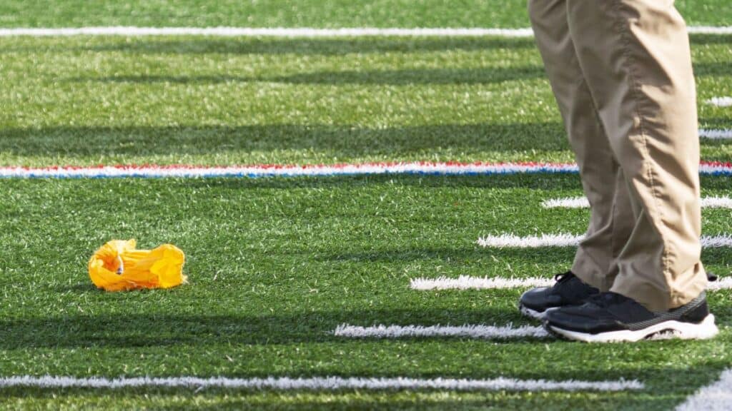 A yellow penalty flag lies on a football field near a person wearing khaki pants and black sneakers—setting the perfect scene for your next fantasy football draft party hosting event.