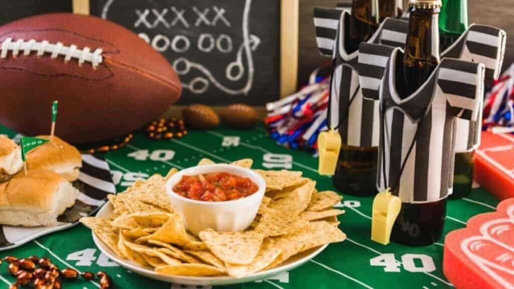 A plate of tortilla chips with salsa, sliders, and beers sits on a football field tablecloth, surrounded by football-themed decorations—perfect for fantasy football draft party hosting.