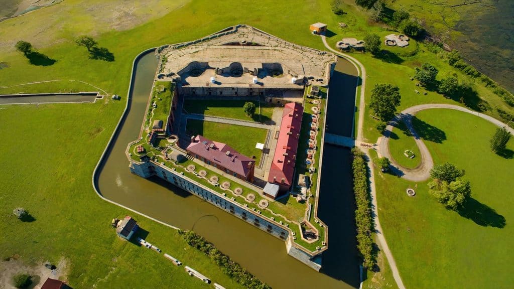 Aerial view of a pentagon-shaped fortress surrounded by a moat, with grassy areas, pathways, and scattered trees in the vicinity.