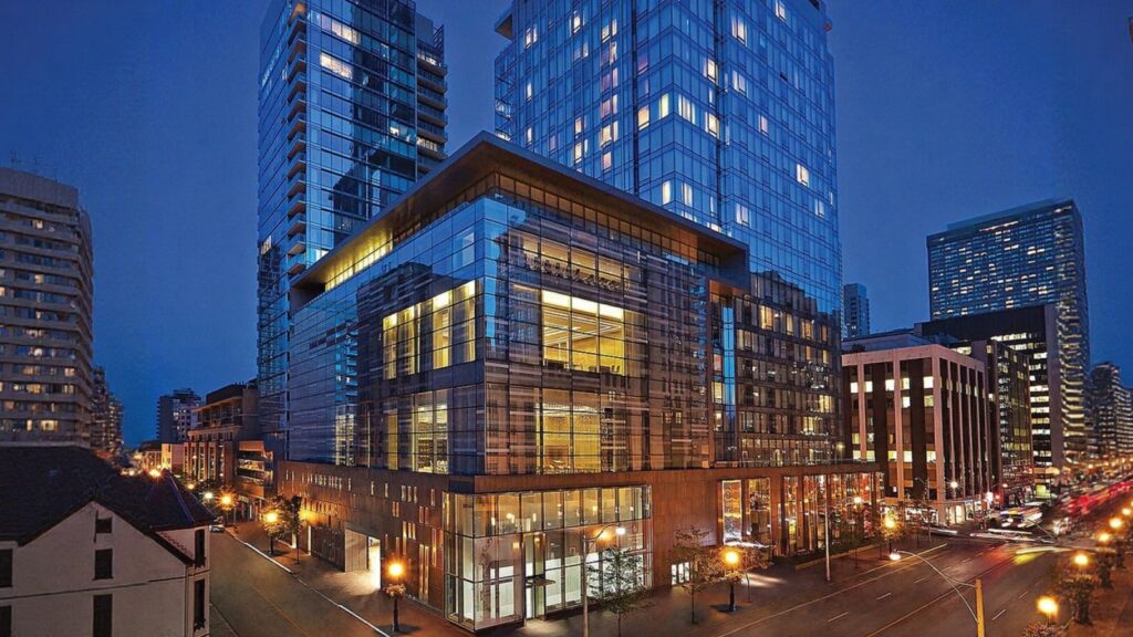 Glass and steel high-rise buildings illuminated at dusk along a busy city street, with cars and streetlights visible.