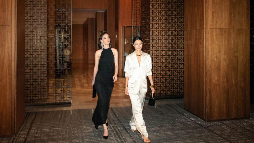 Two women walk through a modern, wood-paneled hallway; one wears a black dress and heels, the other wears a white suit and carries a small purse.