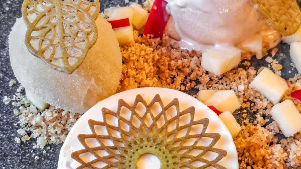 Dessert plate with two spheres, decorative cookie crisps, white sauce, crumbled nuts, and small diced fruit pieces on a dark surface.