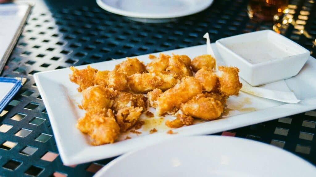 Rectangular white plate with fried, breaded bite-sized Packers cheese curds and a small square dish of white dipping sauce on a black metal table.