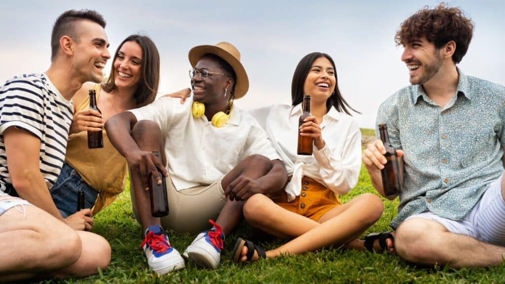 Five young adults sitting on grass, smiling and holding beer bottles, enjoying an outdoor gathering on a sunny day&mdash;thankfully without worrying about steep NFL stadium beer prices.