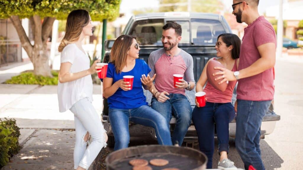 Five people talk and smile while holding red cups, sitting and standing by a truck tailgate, with a grill and hamburgers in the foreground&mdash;capturing the spirit of America&rsquo;s greatest game day tradition.
