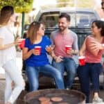 Five people talk and smile while holding red cups, sitting and standing by a truck tailgate, with a grill and hamburgers in the foreground—capturing the spirit of America’s greatest game day tradition.