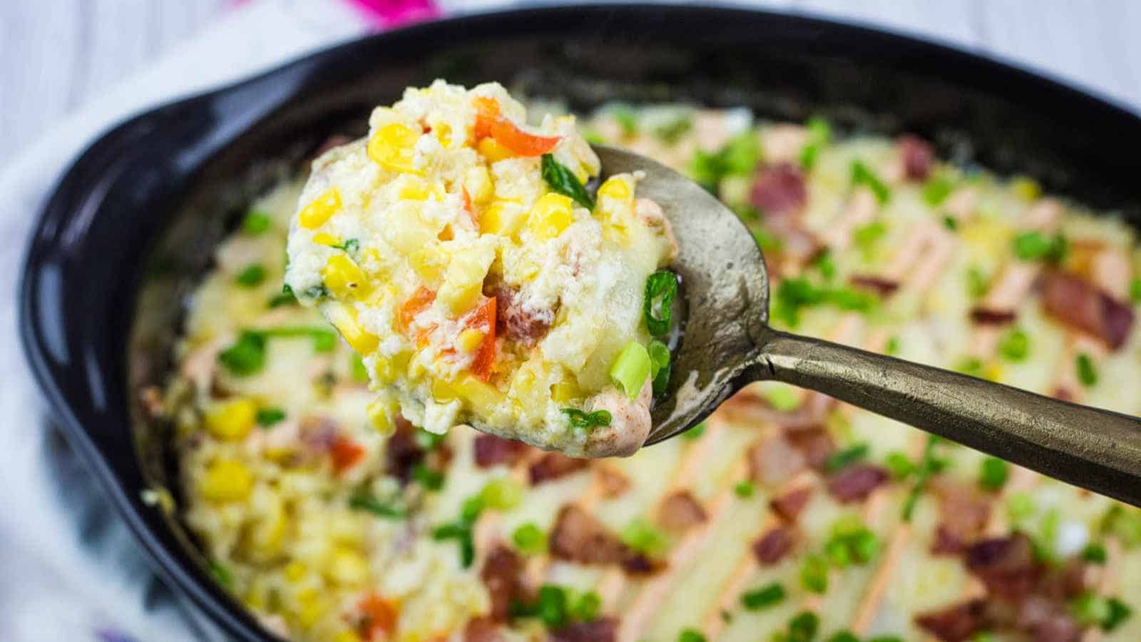 A spoonful of fully loaded corn casserole held above a baking dish of the same casserole.