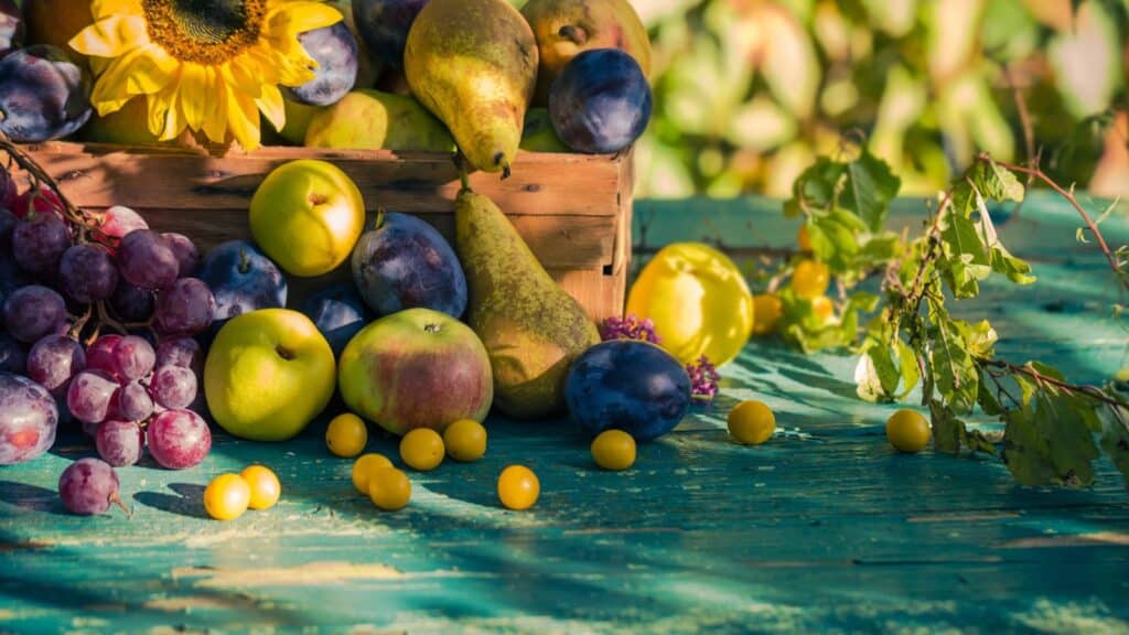 A wooden crate filled with apples, pears, plums, and grapes sits on a rustic blue table with scattered yellow berries and a sunflower.