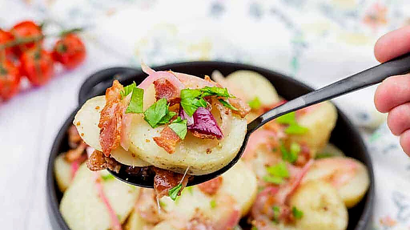 A spoonful of German potato salad topped with bacon, red onion, and chopped parsley held over a bowl of the same salad.