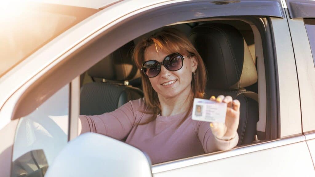 Woman sitting in a car driver&rsquo;s seat, wearing sunglasses, smiling, and proudly holding up her US driver&rsquo;s license toward the camera&mdash;excited to use her US driver&rsquo;s license abroad.