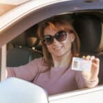 Woman sitting in a car driver’s seat, wearing sunglasses, smiling, and proudly holding up her US driver’s license toward the camera—excited to use her US driver’s license abroad.