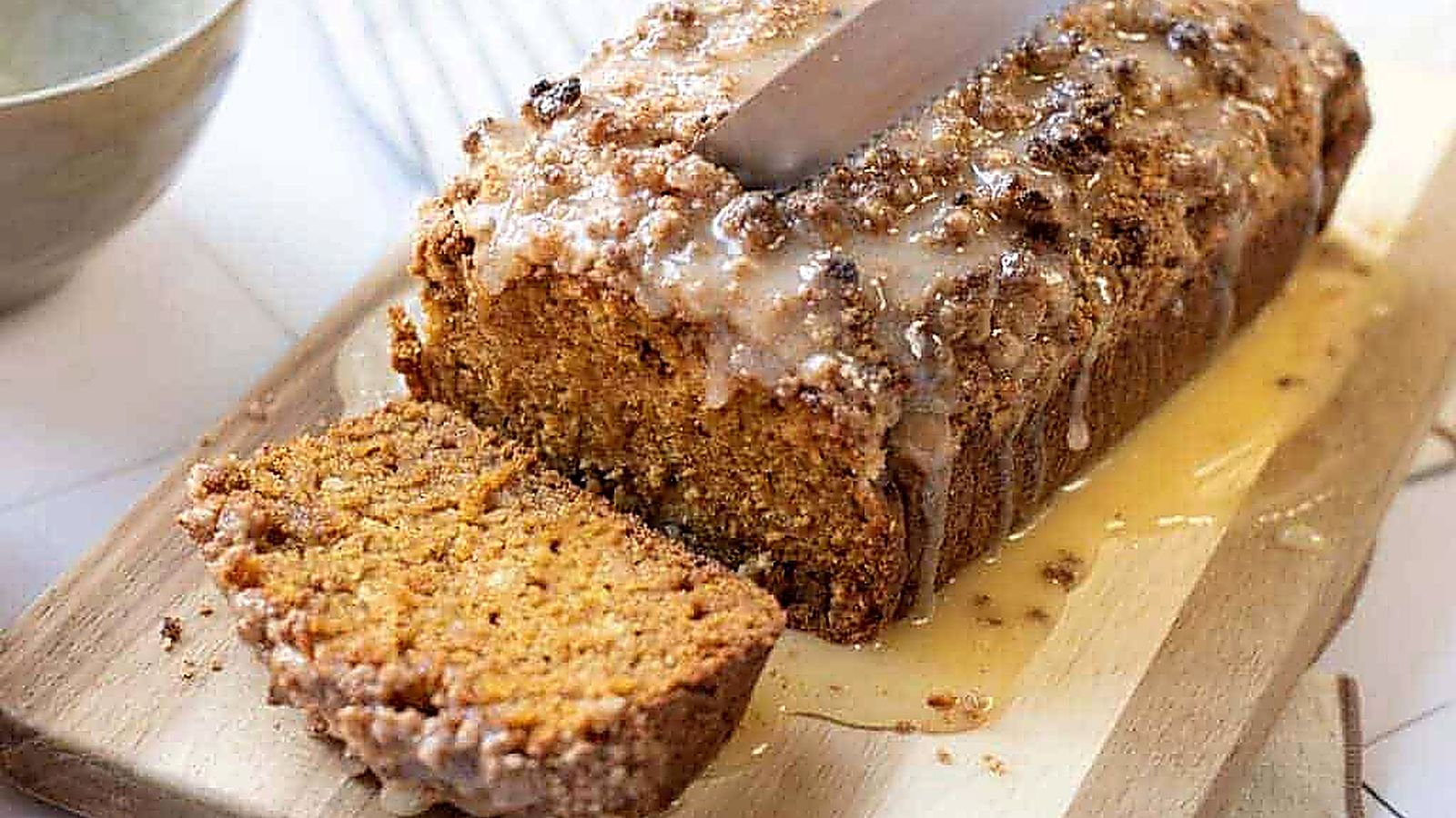A loaf of pumpkin bread with streusel sits on a wooden cutting board; one slice is cut, and a knife rests on top.