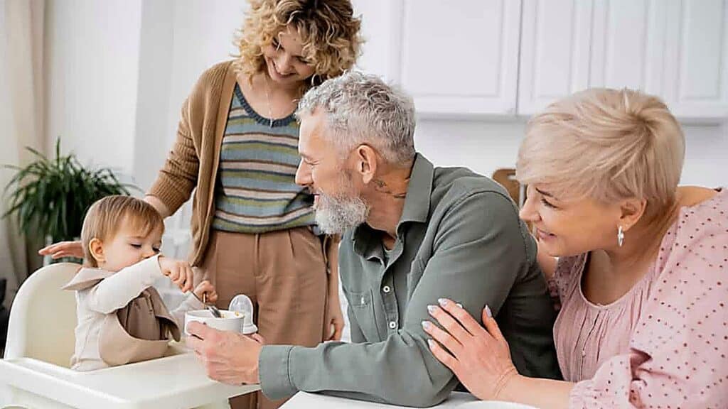 Three adults watch as a toddler in a high chair reaches for food. The group appears to be in a bright kitchen setting.