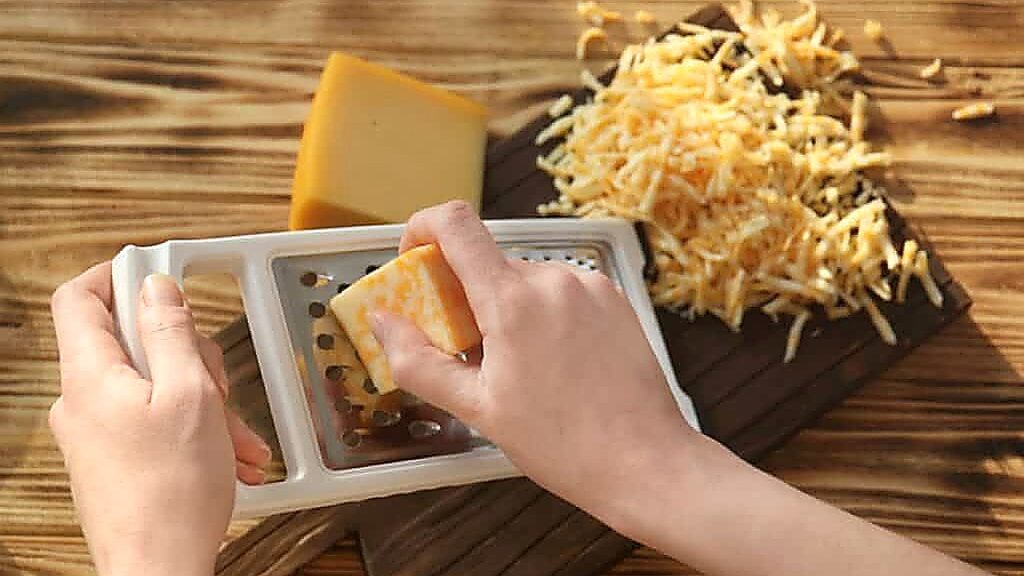 A person grates a block of cheese on a box grater, with shredded cheese and another block of cheese on a wooden surface, showcasing the versatility for ingredient substitutions in your favorite recipes.