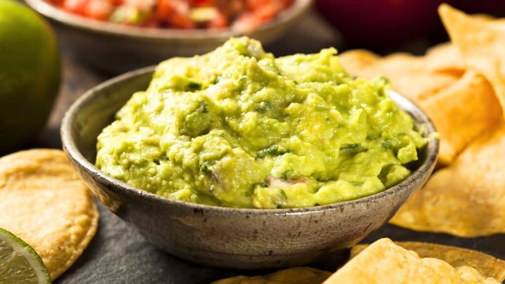 A bowl of guacamole surrounded by tortilla chips, with a lime and salsa blurred in the background.