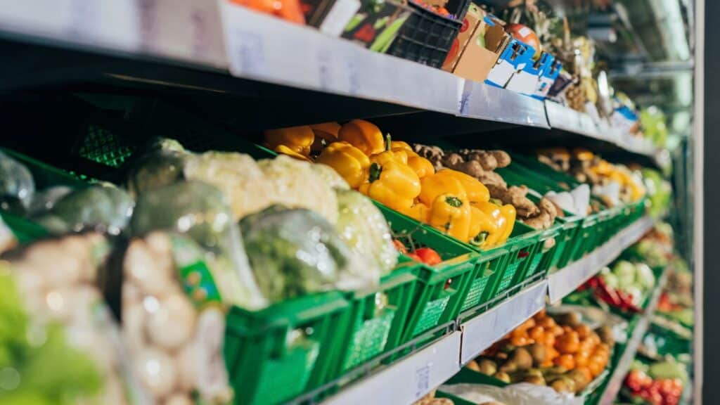 Shelves in a grocery store display green baskets filled with assorted fresh vegetables, including yellow bell peppers, broccoli, and other produce.