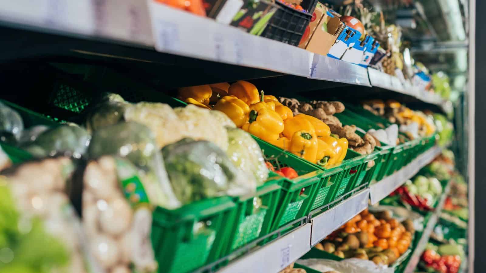 Shelves in a grocery store display green baskets filled with assorted fresh vegetables, including yellow bell peppers, broccoli, and other produce.