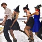 Four people in witch costumes and one in a hat pose on two broomsticks in a park, smiling and playfully pretending to fly.