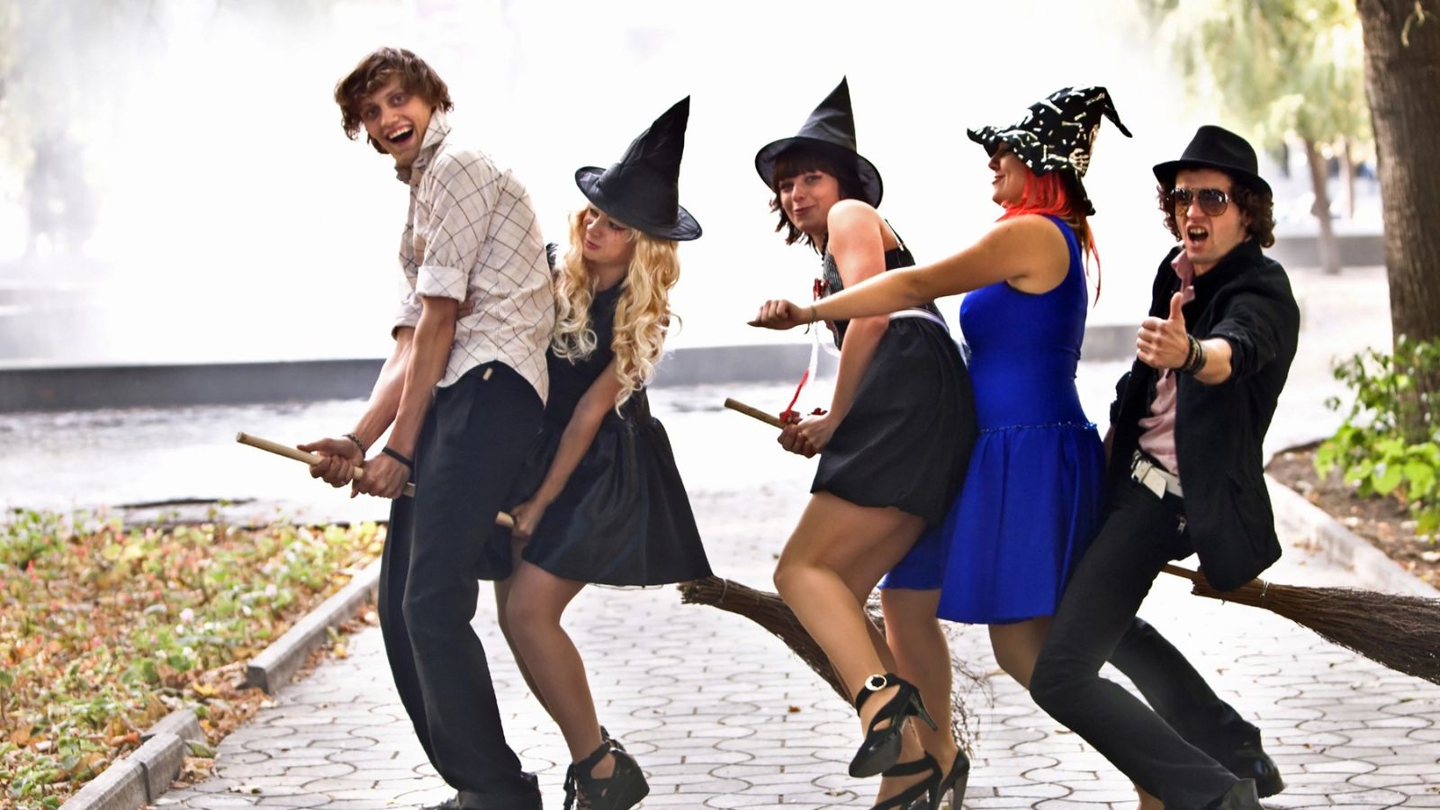 Four people in witch costumes and one in a hat pose on two broomsticks in a park, smiling and playfully pretending to fly.