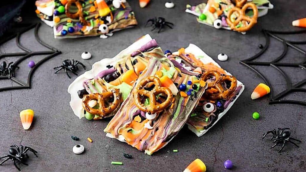 Pieces of white chocolate bark decorated with pretzels, candy corn, and candy eyeballs, scattered with plastic spider decorations.
