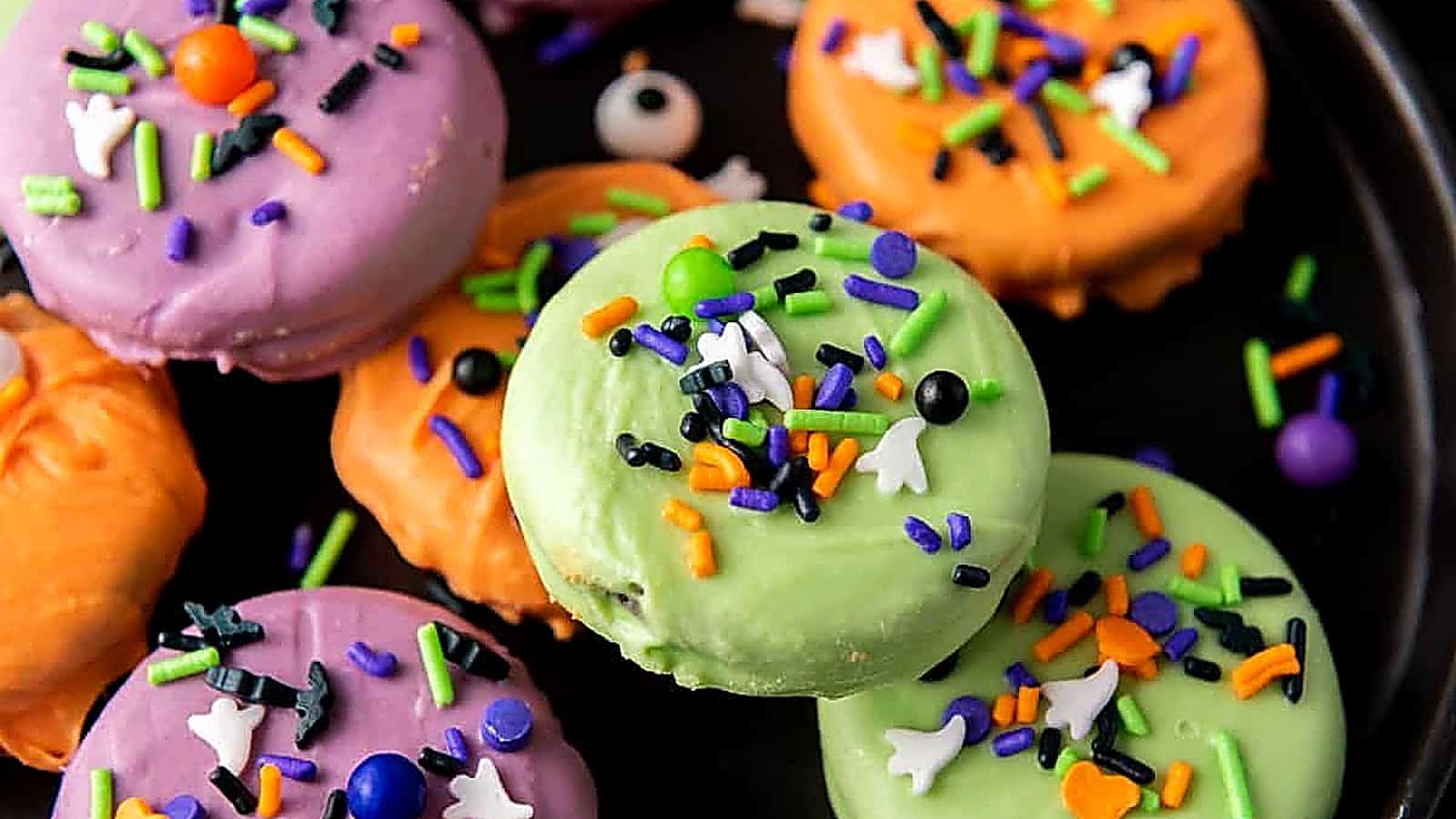 A group of Halloween Oreo Cookies coated in green, orange, and purple, decorated with sprinkles and candy eyes, sits on a gray surface next to a black cloth.
