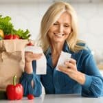 A woman in a kitchen looks at a receipt while sitting near a paper bag filled with fresh groceries including lettuce, bread, and vegetables.
