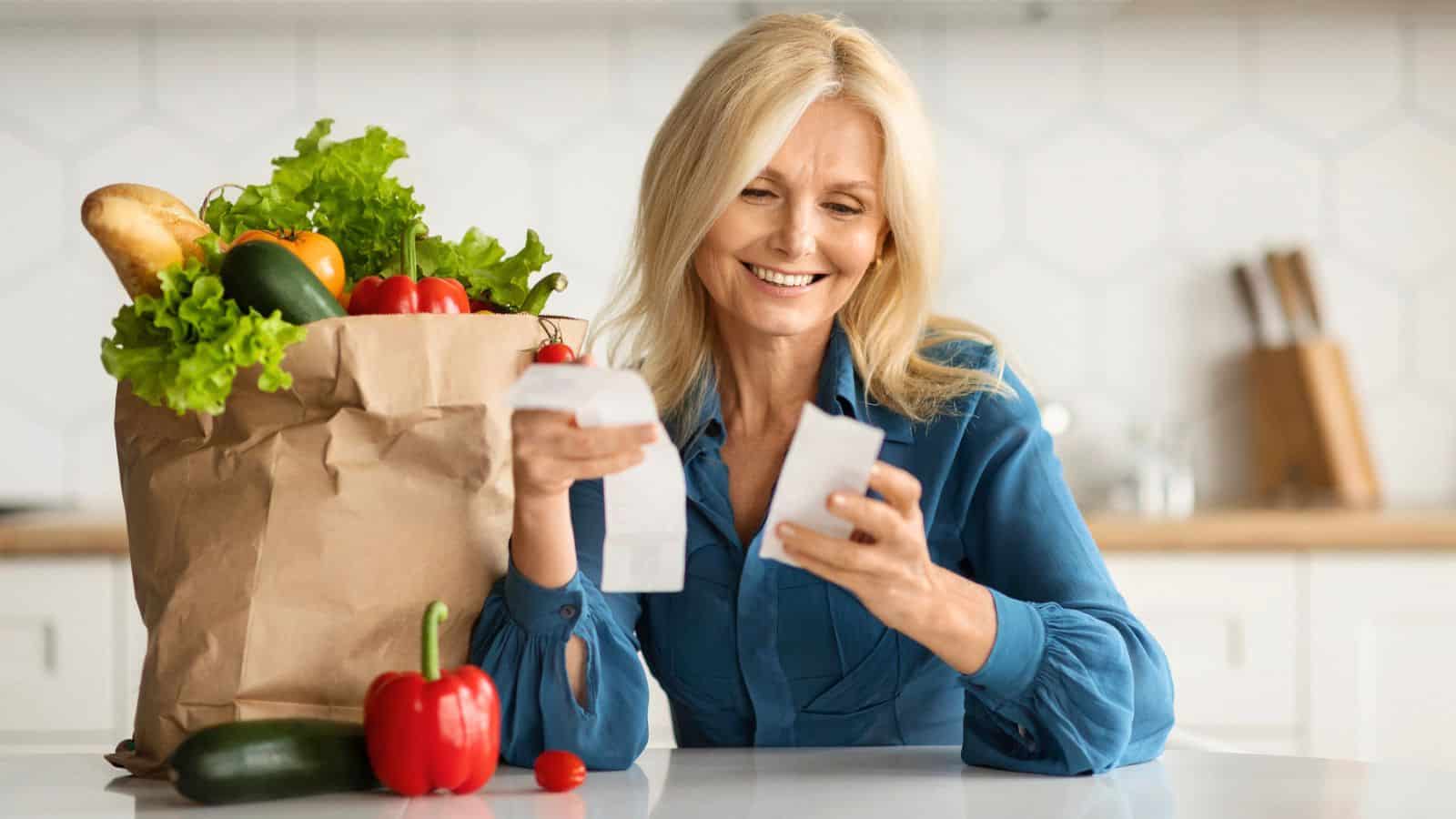 A woman in a kitchen looks at a receipt while sitting near a paper bag filled with fresh groceries including lettuce, bread, and vegetables.