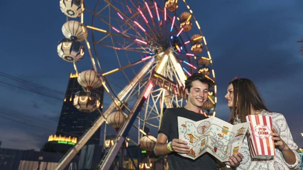 Two people stand in front of a lit Ferris wheel at night, holding a map and a popcorn container, smiling and enjoying the carnival atmosphere&mdash;all while noting the importance of following Disney and Universal theme park rules for a safe and memorable experience.