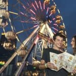 Two people stand in front of a lit Ferris wheel at night, holding a map and a popcorn container, smiling and enjoying the carnival atmosphere—all while noting the importance of following Disney and Universal theme park rules for a safe and memorable experience.