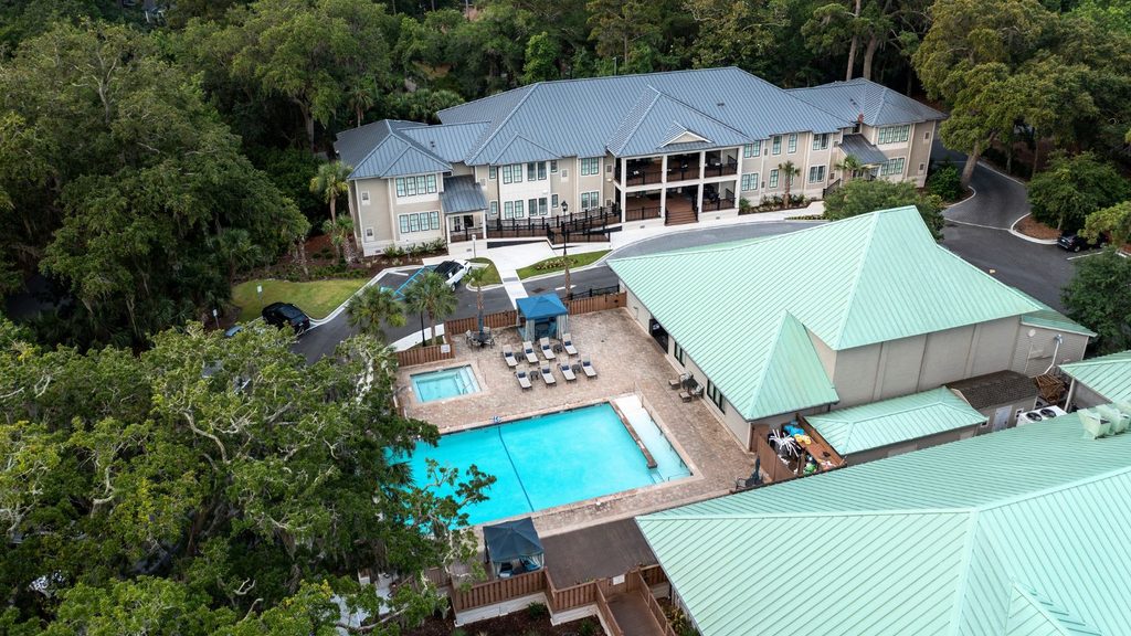 Aerial view of a residential complex with a large outdoor swimming pool, lounge chairs, umbrellas, and surrounding trees. Two main buildings with green roofs are visible, designed to offer a relaxing environment that also features wellness amenities for glp-1 support.