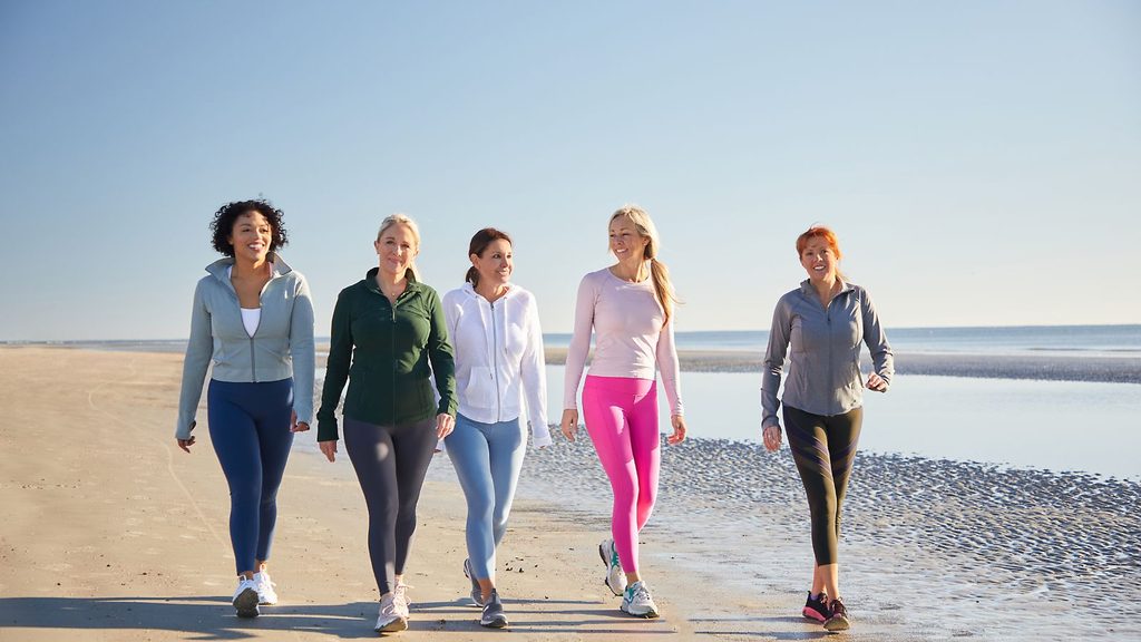 Five women walk together on a sandy beach in athletic wear under a clear sky, with water and shoreline visible in the background, embodying the spirit of wellness and glp-1 support.